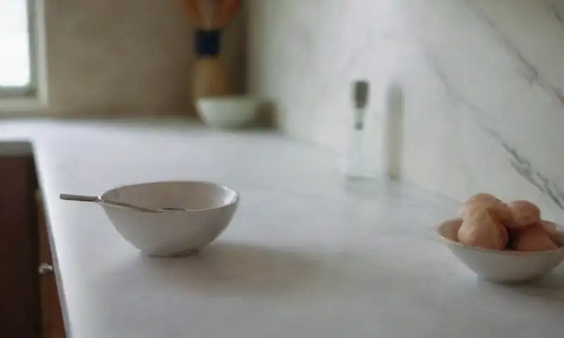 Un elegante mostrador de mármol blanco brilla suavemente bajo la luz natural, con elementos como un bol de bicarbonato y utensilios, creando un ambiente acogedor en una cocina ordenada