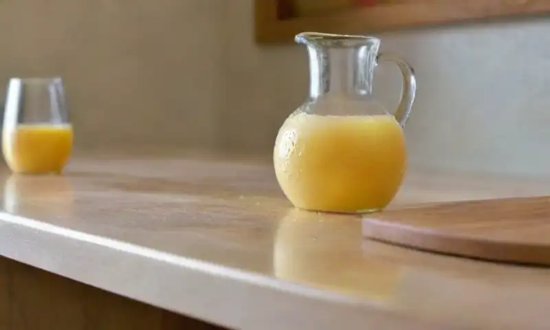 Una cocina bien iluminada muestra un encimera pulida con una jarra de café, mezcla de vinagre y agua, un limón cortado, una esponja, una tabla de madera, una receta abierta y granos de café aromáticos dispersos, creando un ambiente acogedor