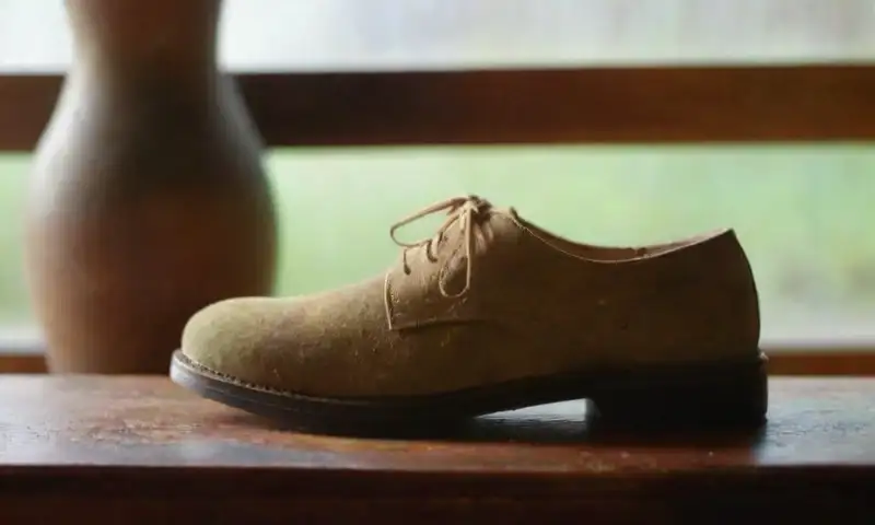 Unas suaves botas de ante descansan sobre una mesa de madera rústica, iluminadas por la luz natural que resalta sus detalles y una botella de limpiador cercana
