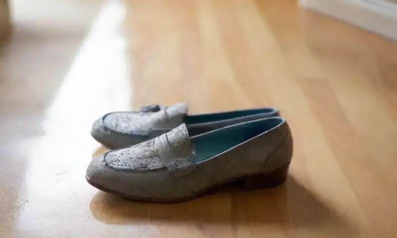 Una habitación iluminada con rayos de sol, donde unos zapatos de gamuza taupe descansan sobre una tela blanca, junto a un tazón de agua y un cepillo de dientes desgastado, reflejando un proceso de limpieza cuidadoso