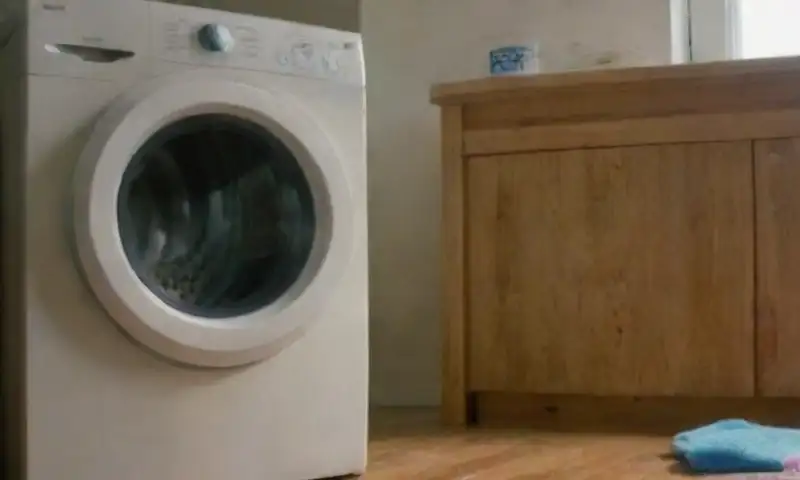 Una lavadora de carga frontal brilla con luz ambiental, mostrando ropa mojada y un panel de control detallado, junto a botellas de detergente y un suelo de madera cálida